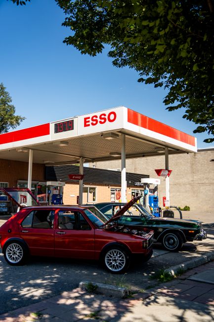 A bright, sunny day at a petrol station with an ESSO canopy displaying an electronic price sign showing fuel prices. Two cars are parked at the pumps: a red hatchback with a slightly open bonnet and a