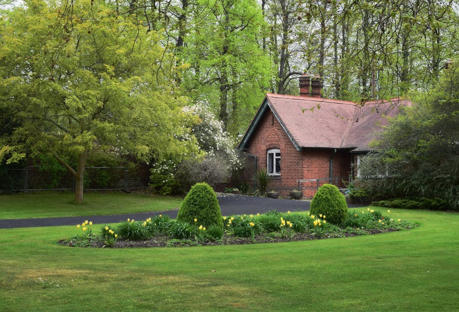 A well-maintained front garden featuring lush green grass with several flowerbeds bordered by dark, rich soil. The flowerbeds are filled with vibrant pink and white flowers, arranged in neat, curved l