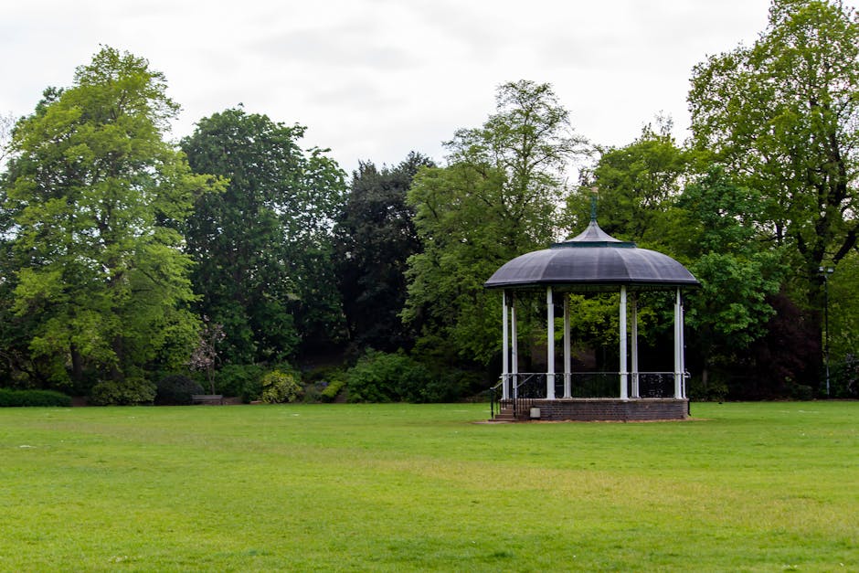 A peaceful park scene featuring a circular bandstand with a dark, domed roof supported by white columns, situated on a small brick platform. Surrounding the bandstand is a well-maintained, lush green 